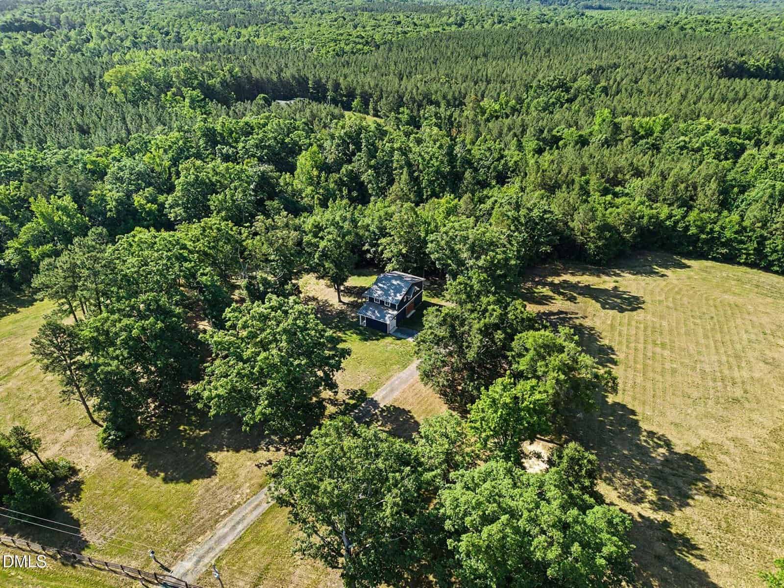 Aerial view of a small house at 8196 Raymond Royster Road, Roxboro, surrounded by dense trees and greenery, with a driveway leading to the house, open grassy areas, and a forest stretching into the distance under a clear sky.