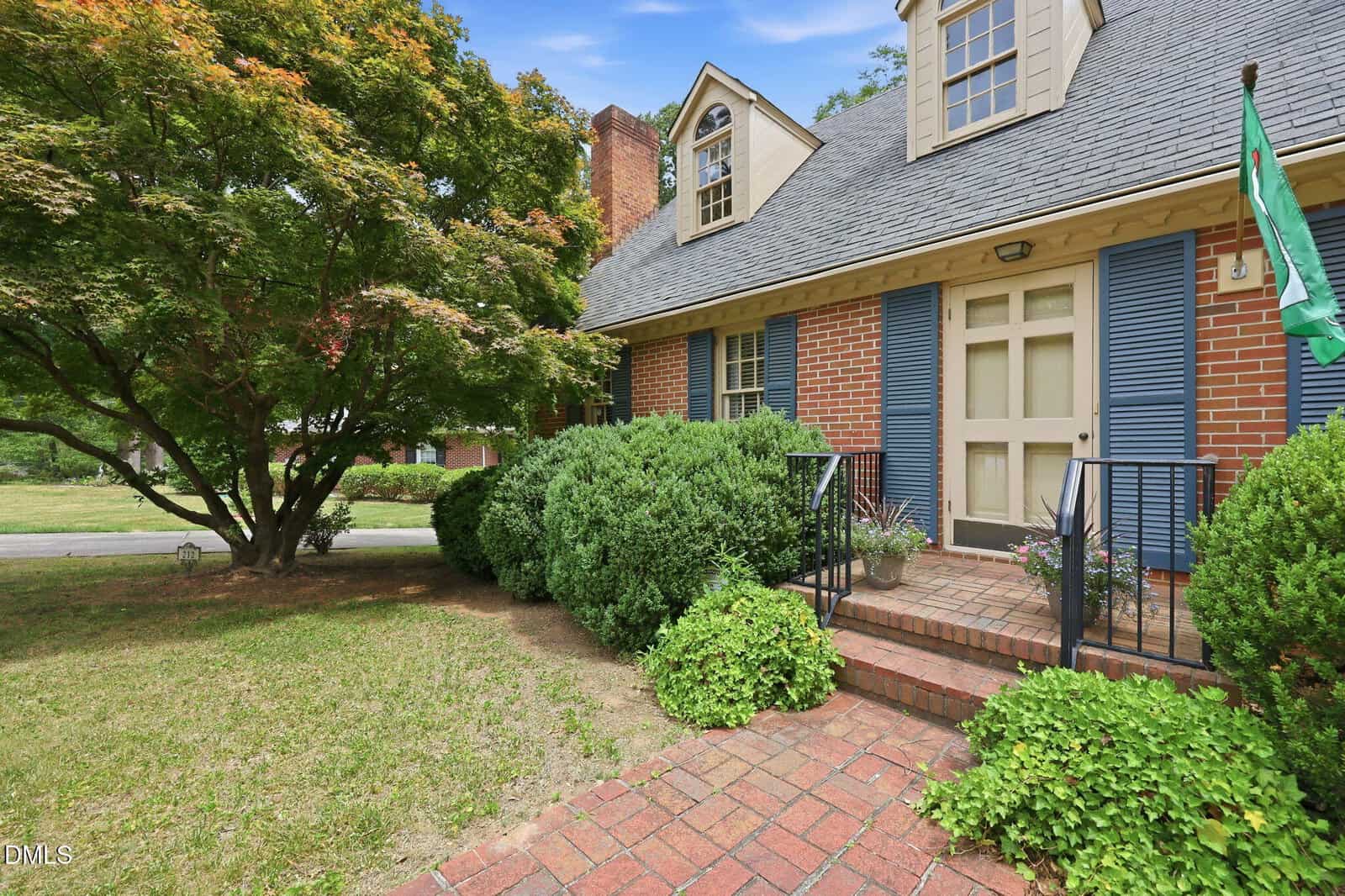 A charming brick house at 212 Hillcrest Drive in Oxford, with blue shutters and a gabled roof, features a front porch with black railings, lush shrubs along the walkway, and a mature tree in the yard under a blue sky.