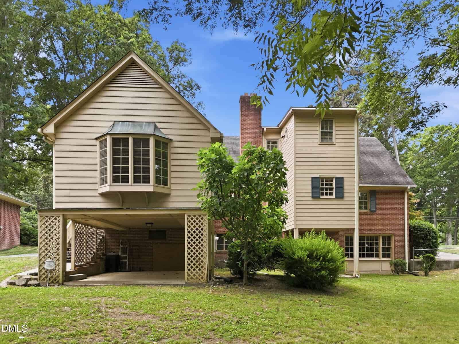 A two-story house with beige siding and red brick exterior, featuring a covered carport, a bay window, green shutters, and surrounding trees and bushes on a grassy lawn under a blue sky.