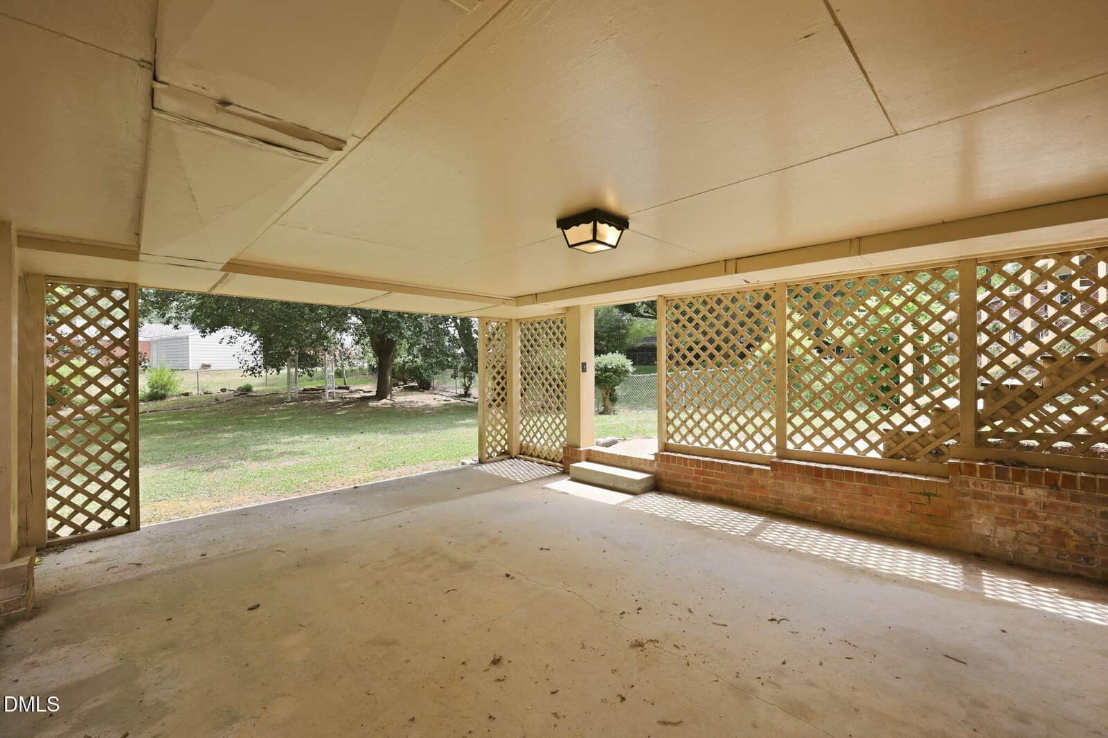 Covered patio with a cement floor, lattice privacy panels on two sides, a brick half-wall, and steps leading to a grassy backyard with trees. Natural light comes through the open areas and the ceiling light fixture.
