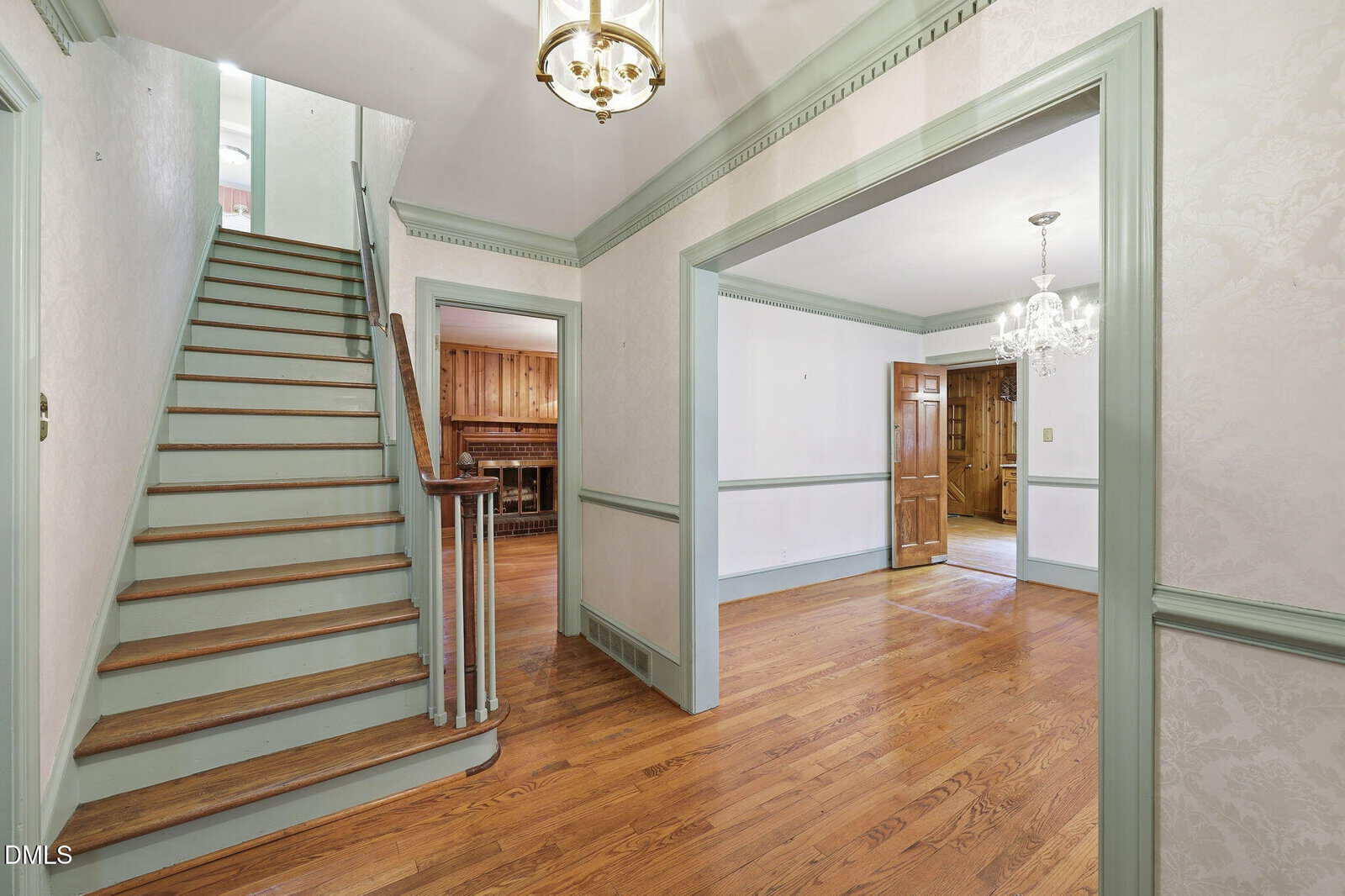 A hallway with hardwood floors features a staircase with wooden steps and light green trim, leading upstairs. An open doorway reveals a dining room with a chandelier and wainscoting, and another room with wood-paneled walls.
