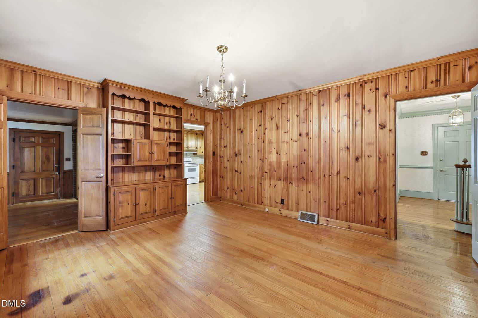 A wood-paneled room with built-in shelves, chandelier, hardwood floors, and open doorways leading to a kitchen and hallway.