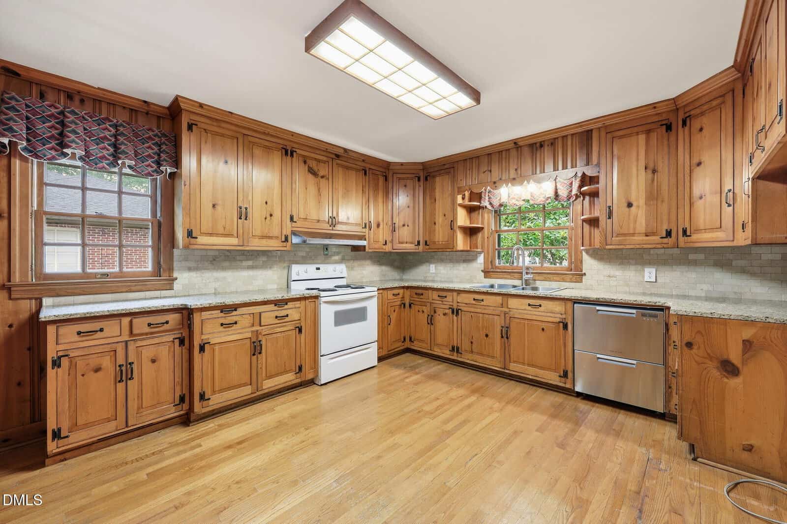 Spacious kitchen with wooden cabinets, hardwood floors, white stove, stainless steel dishwasher, and large windows with valances letting in natural light.