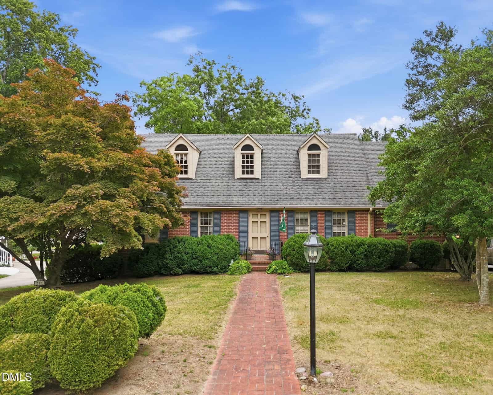 A brick house with three dormer windows, surrounded by trees and bushes, features a red brick walkway leading to the front entrance under a blue sky.