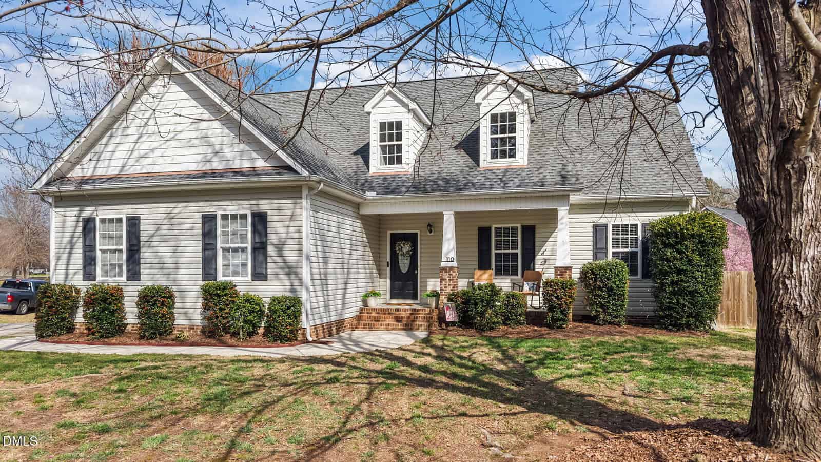 A light gray, single-story house at 110 Willow Ridge Drive in Louisburg features dormer windows, black shutters, a covered front porch, and neatly trimmed bushes, set in a yard with patchy grass and leafless trees under a blue sky.