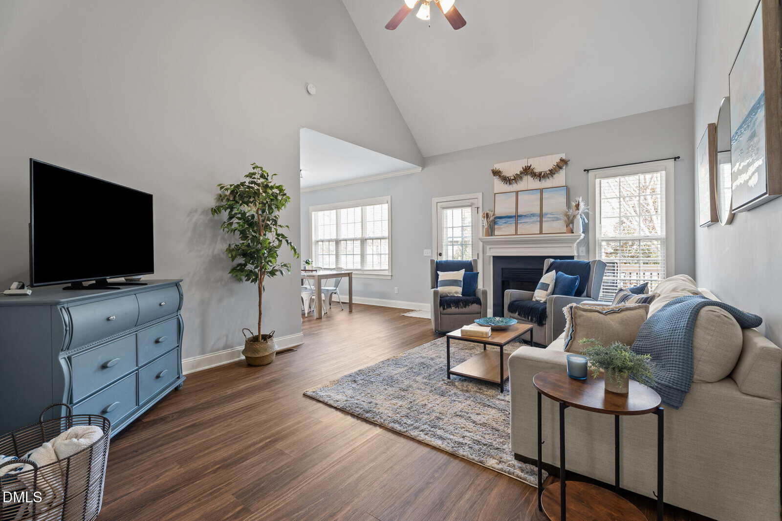 A bright living room with wood floors, a beige sofa, a blue dresser with a TV, a fireplace, plants, and large windows letting in natural light. A dining area is visible in the background.