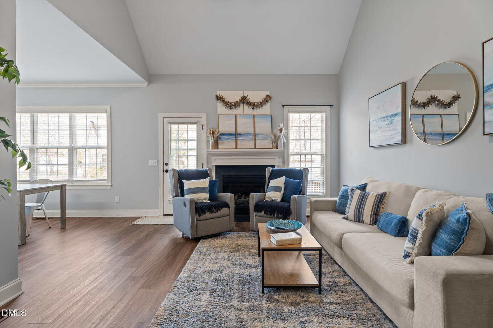 Bright living room with a beige sofa, blue and beige pillows, a patterned rug, two armchairs by a fireplace, and wall art. Large windows let in natural light; an office area is visible in the background.