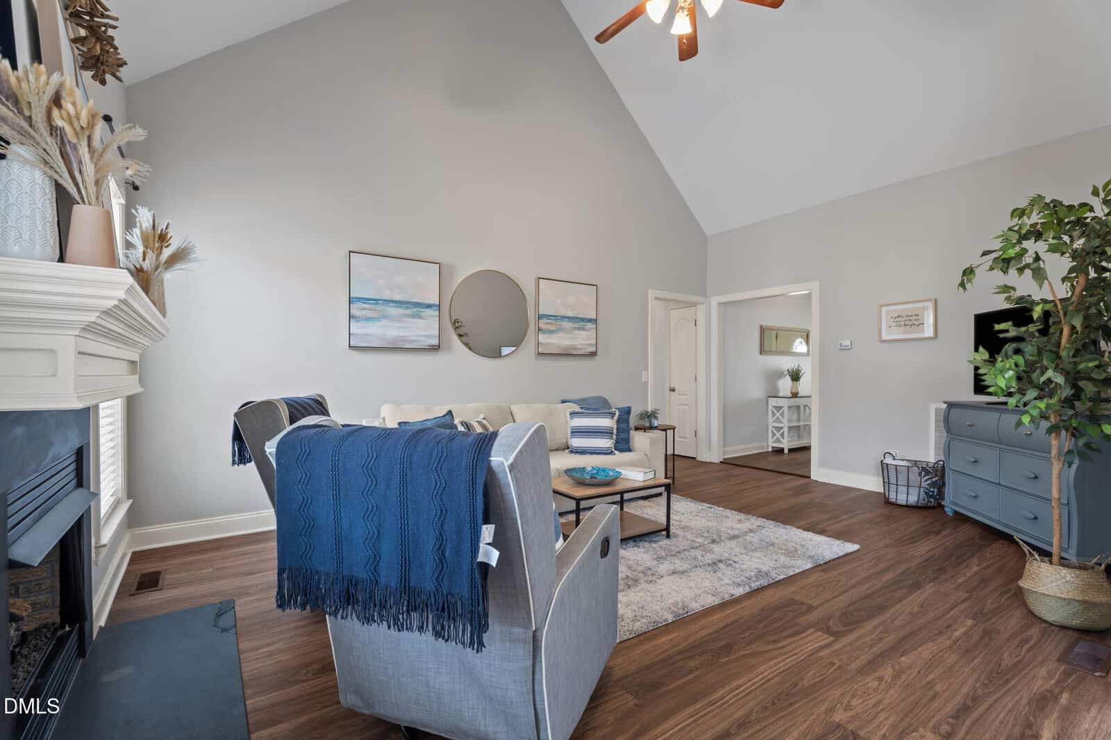 A cozy living room with vaulted ceiling, a ceiling fan, neutral walls, wood floors, a gray sofa, blue armchair, coffee table, fireplace, wall art, a round mirror, TV on a dresser, and a potted tree in the corner.