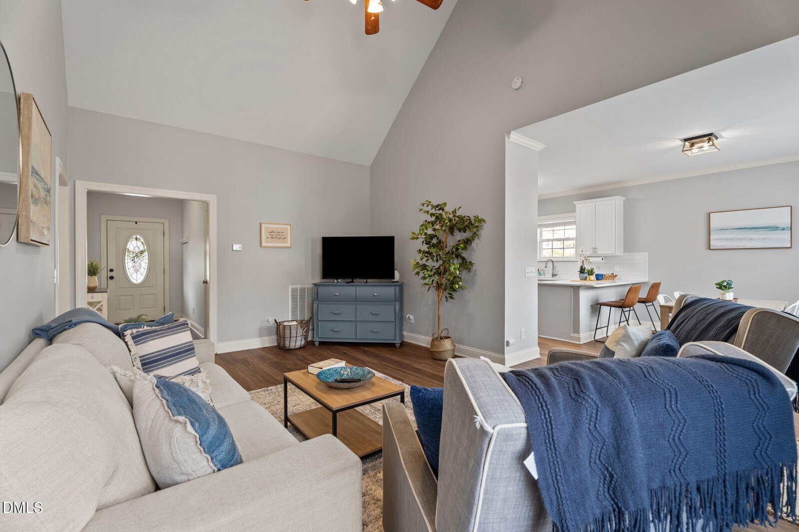 Modern living room with neutral sofas, blue accents, TV on a dresser, potted plant, and view into a bright kitchen and dining area. Walls are light gray, and wood floors add warmth.