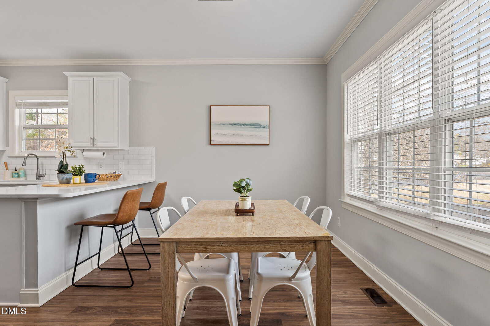 Bright kitchen and dining area with a wooden table, white chairs, breakfast bar with brown stools, large window with blinds, white cabinets, and minimalist wall art.