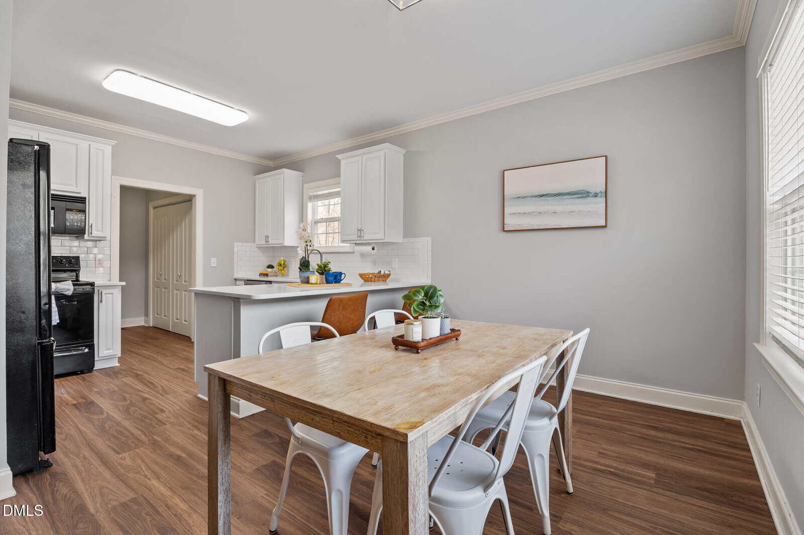 Bright kitchen and dining area with wood floors, light gray walls, white cabinets, a wooden table with white chairs, and a small potted plant centerpiece. A beach painting hangs on the wall above the table.