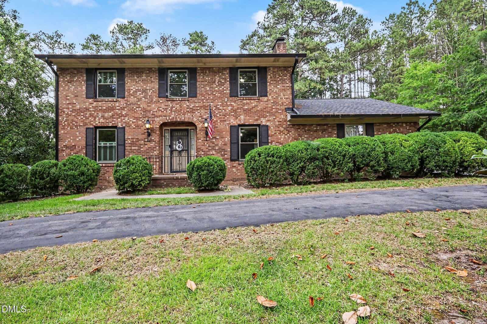 Elegant two-story brick home at 2555 Nutbush Road, Henderson, featuring black shutters, a central front door with an American flag, neatly trimmed bushes, a curved driveway, lush green lawn, and tall trees in the background.