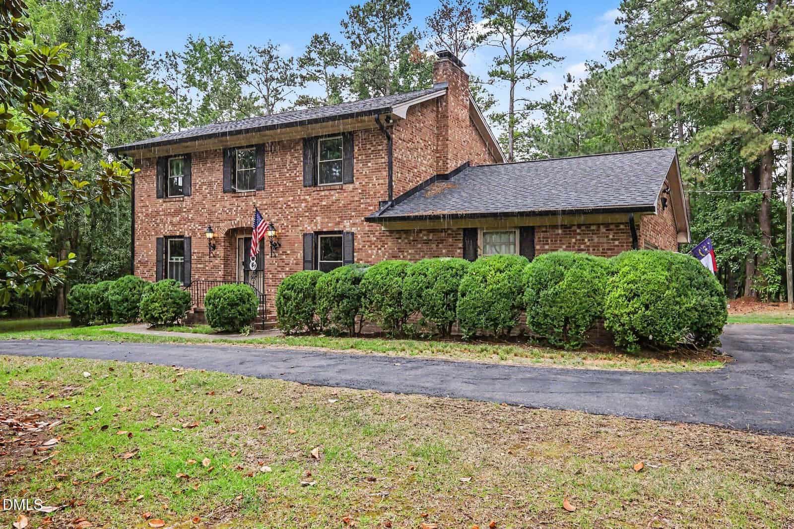 Two-story brick house with black shutters and a front porch, surrounded by neatly trimmed bushes and trees. A curved driveway leads to the entrance, and two flags are displayed near the porch.