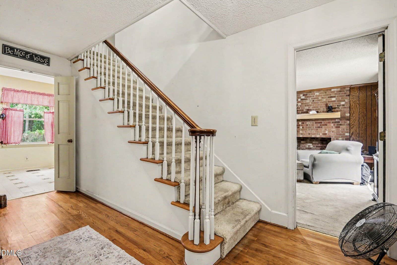 A carpeted staircase with white railings leads upward in a bright hallway with hardwood floors. An open doorway reveals a cozy living room with a brick wall, gray furniture, and a black floor fan.