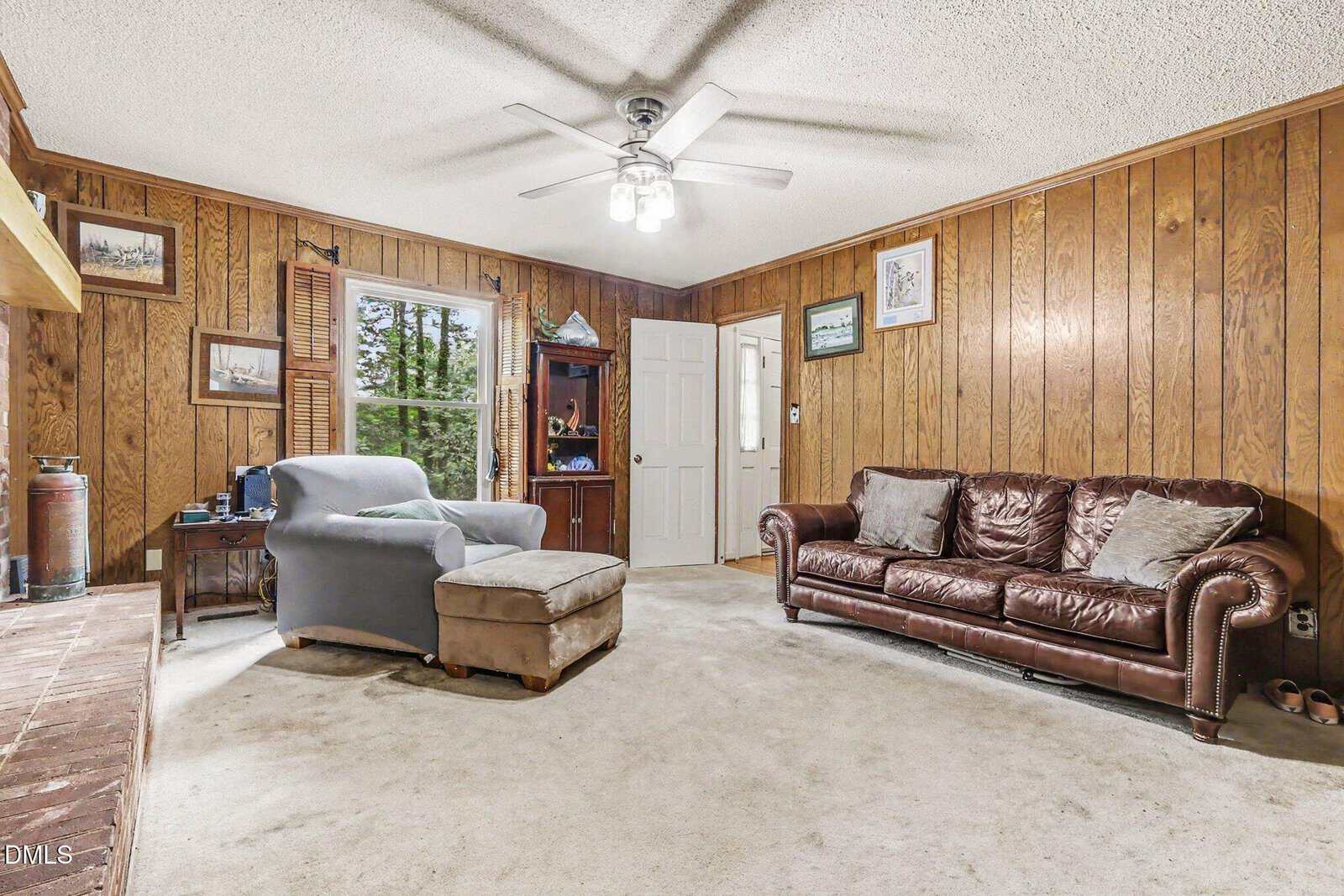 Cozy living room with wood-paneled walls, brown leather sofa, armchair with ottoman, desk, bookshelf, ceiling fan, and window letting in natural light. Carpeted floor and various framed pictures on the walls.