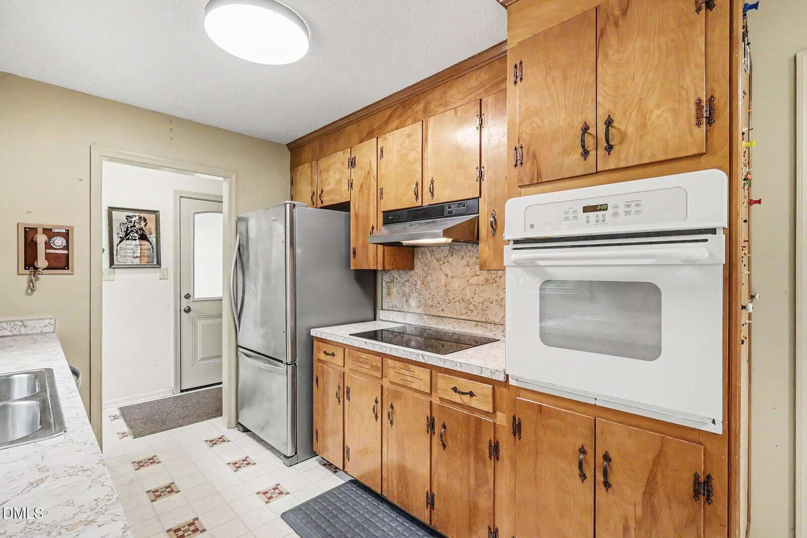 Narrow kitchen with wood cabinets, white wall oven, stainless steel refrigerator, electric cooktop, and marble backsplash. There’s a sink on the left and a door at the back. The floor has a white and floral tile pattern.
