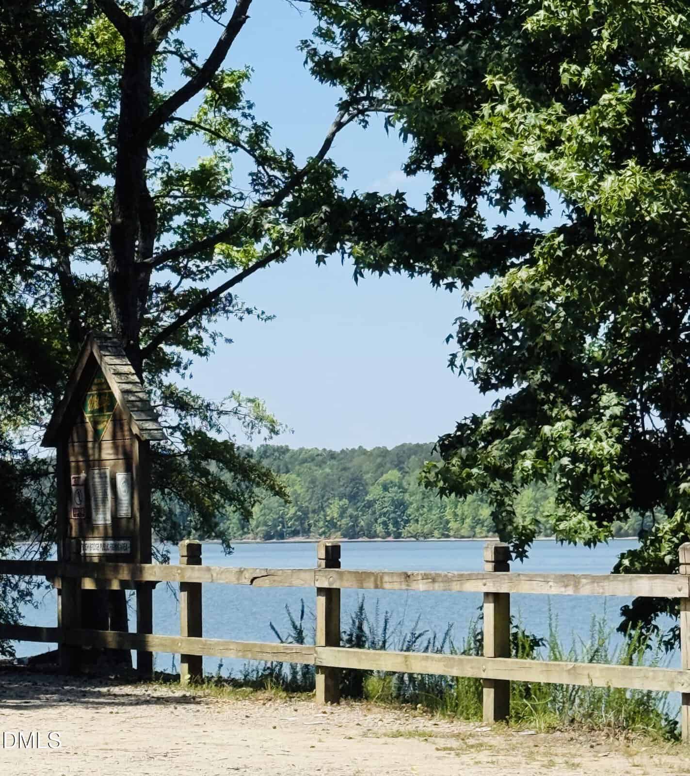 A wooden fence runs along the edge of a lake, with trees providing shade. A wooden signboard is on the left, and forested land is visible across the water under a clear, blue sky.