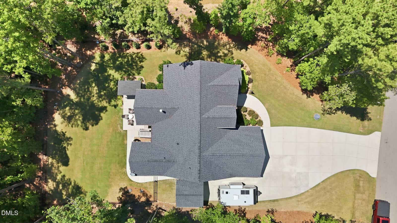 Aerial view of a large house with a dark gray roof, surrounded by trees and a grassy yard. The home features a spacious driveway, backyard patio, and landscaped lawn. The property borders a road on one side.