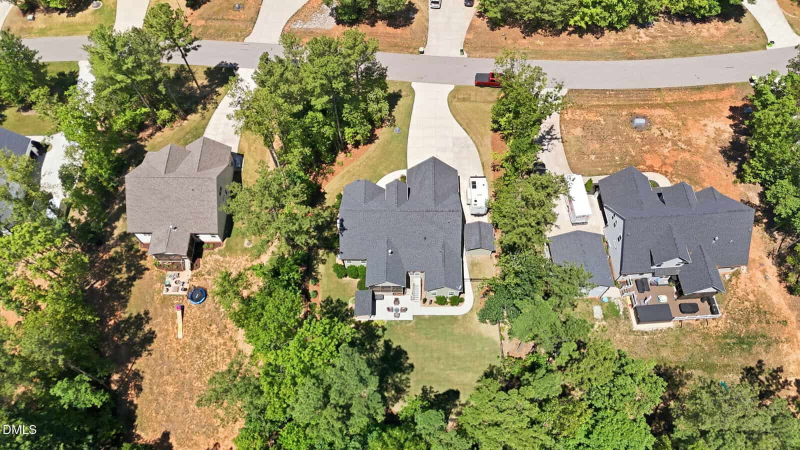 Aerial view of three suburban homes with large backyards, surrounded by trees and greenery, situated along a curved residential street with driveways and a few parked vehicles.