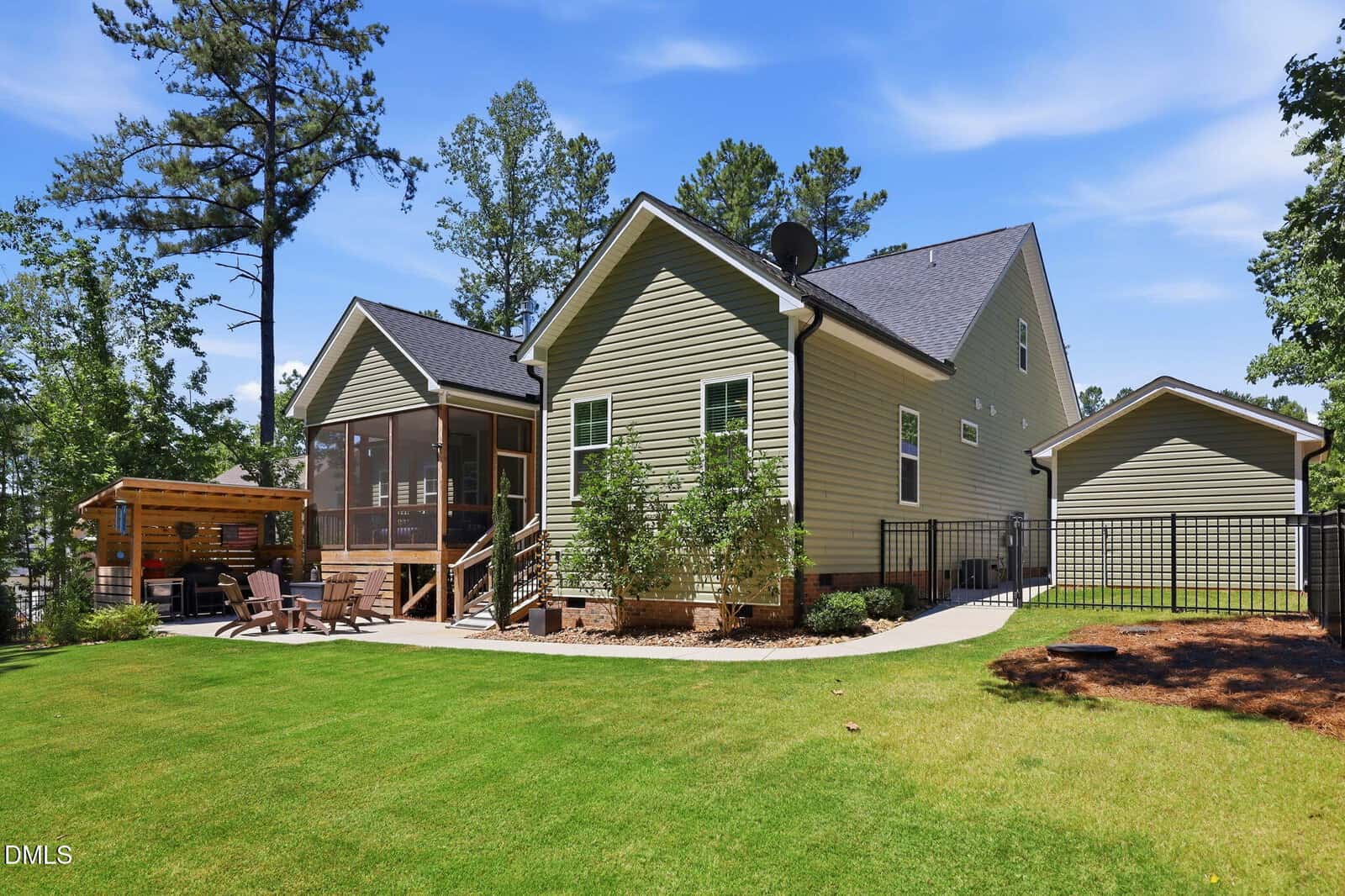 A modern house with light green siding, a screened porch, and a pergola seating area in a spacious backyard with green grass, trees, and a detached garage.