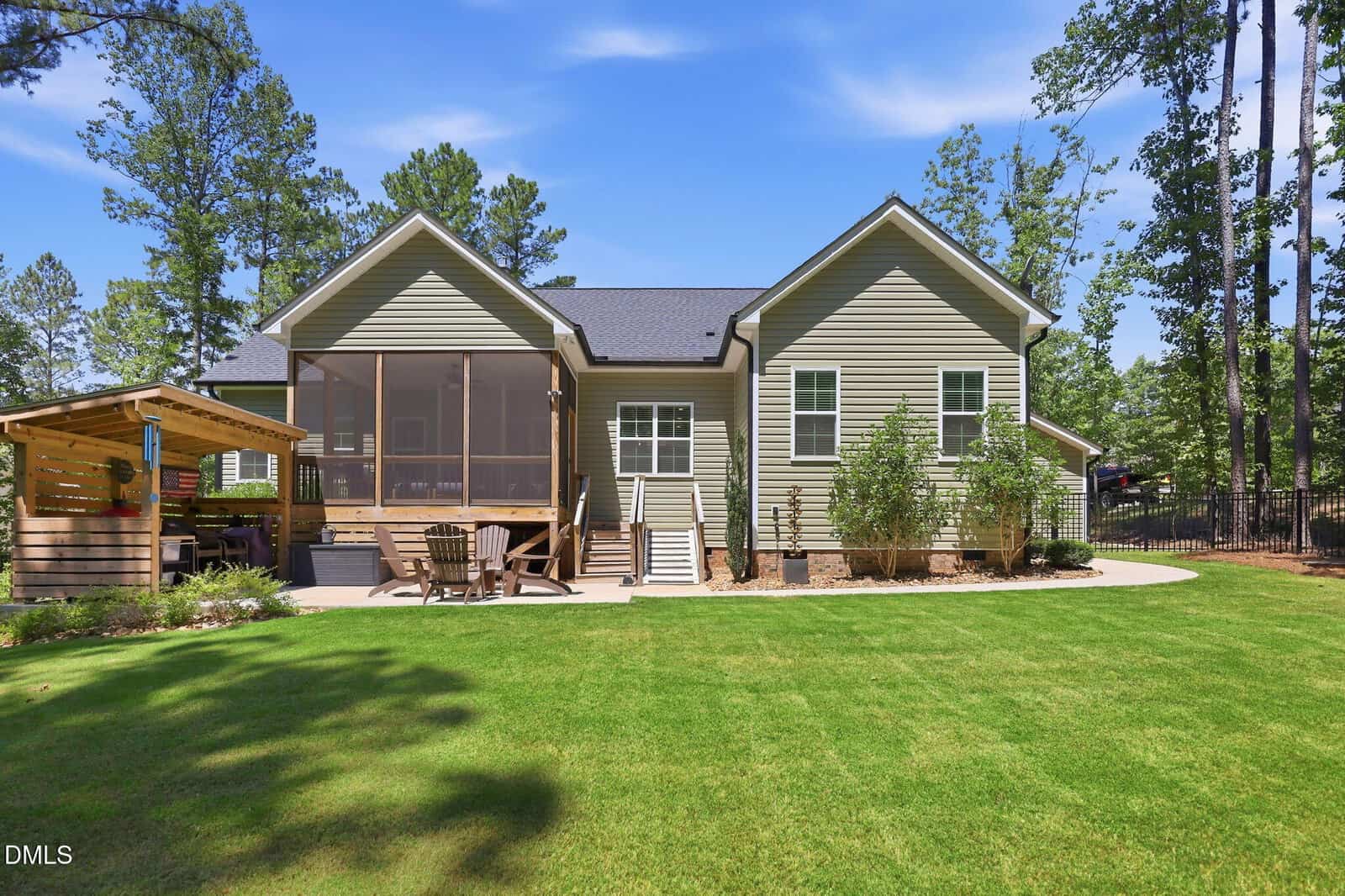 A modern house with light green siding, a screened-in porch, wooden deck, and pergola in a spacious backyard with lush grass, trees, and outdoor seating under a clear blue sky.