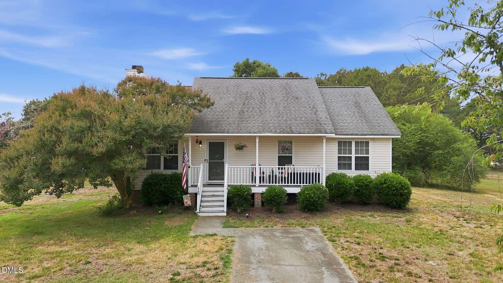 A small white house at 15 Magnolia Ridge Drive, Louisburg, features a front porch with steps and railing, partially shaded by a large tree. Sparse grass and bushes fill the yard, while an American flag hangs by the doorway under a blue sky.