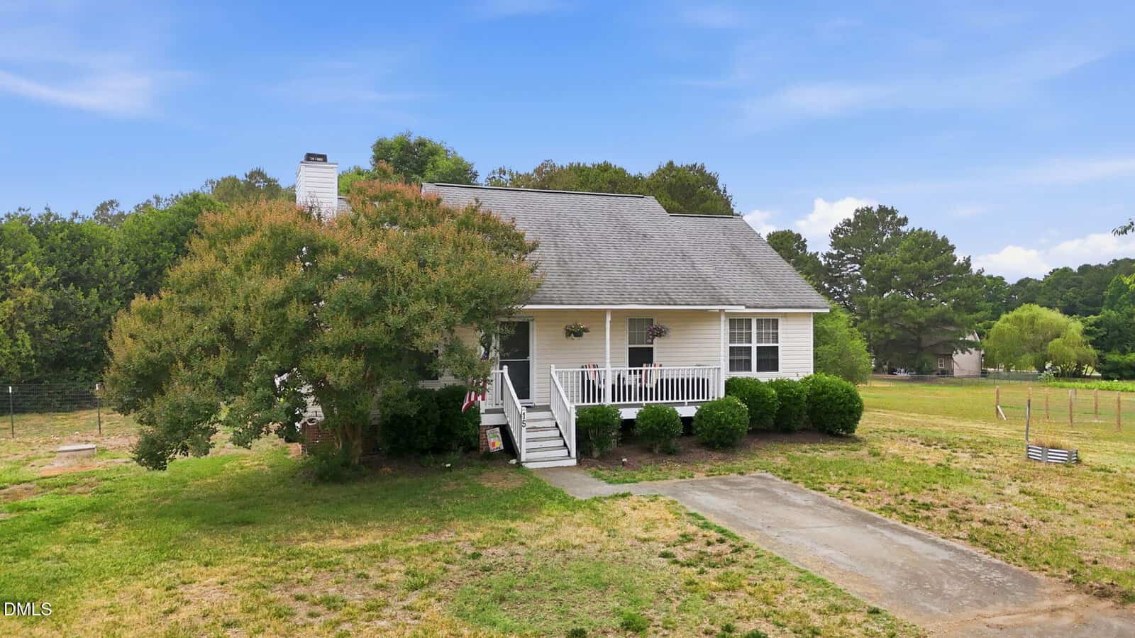 A small white house with a covered front porch and white railing, surrounded by green bushes and trees, sits on a grassy lot with a patchy lawn and a concrete driveway under a blue sky.