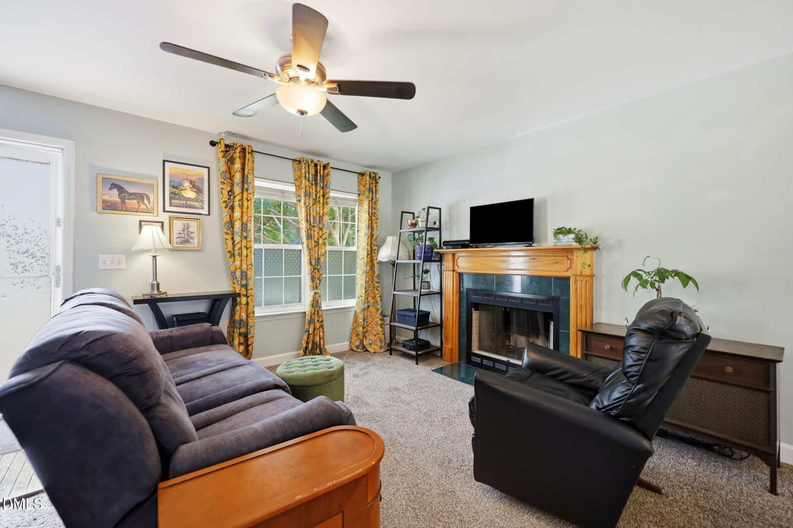 A cozy living room with a blue sofa, black recliner, fireplace with wooden mantel, TV, desk, and two windows with yellow patterned curtains. Carpeted floor and light walls with framed pictures complete the space.