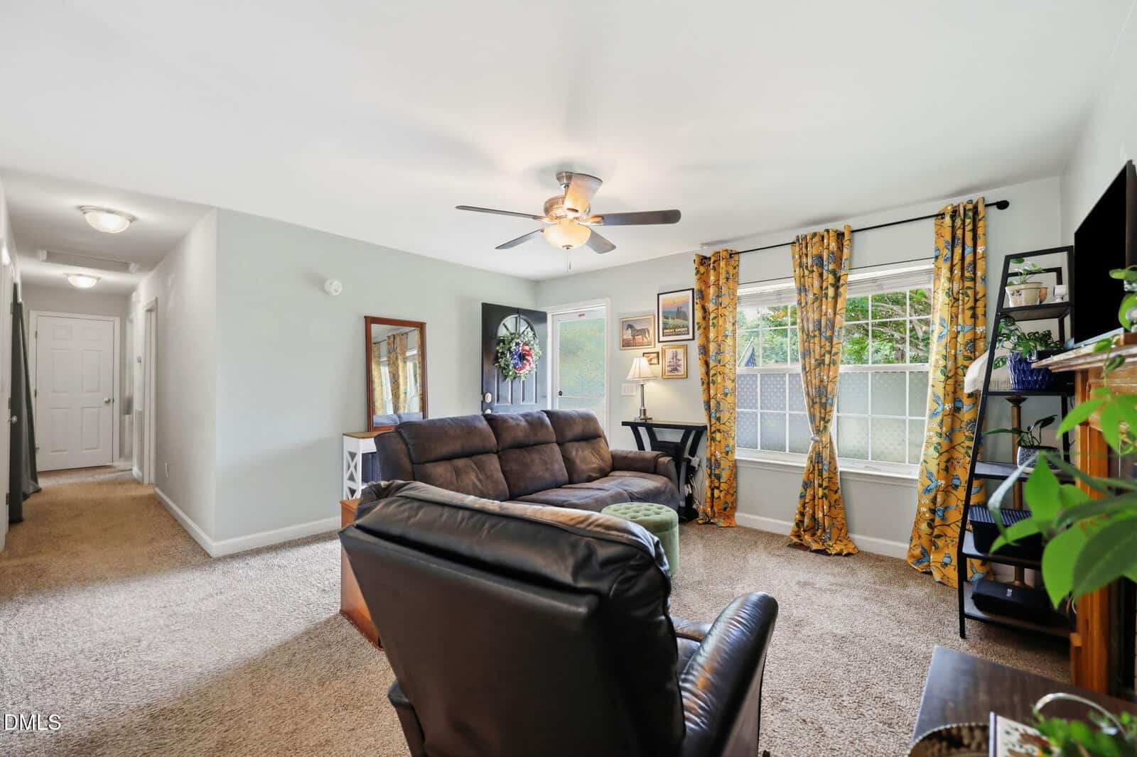 A cozy living room with brown sofas, a ceiling fan, and a TV. Large windows with yellow patterned curtains let in natural light. A hallway leads to other rooms, and various plants and pictures decorate the space.