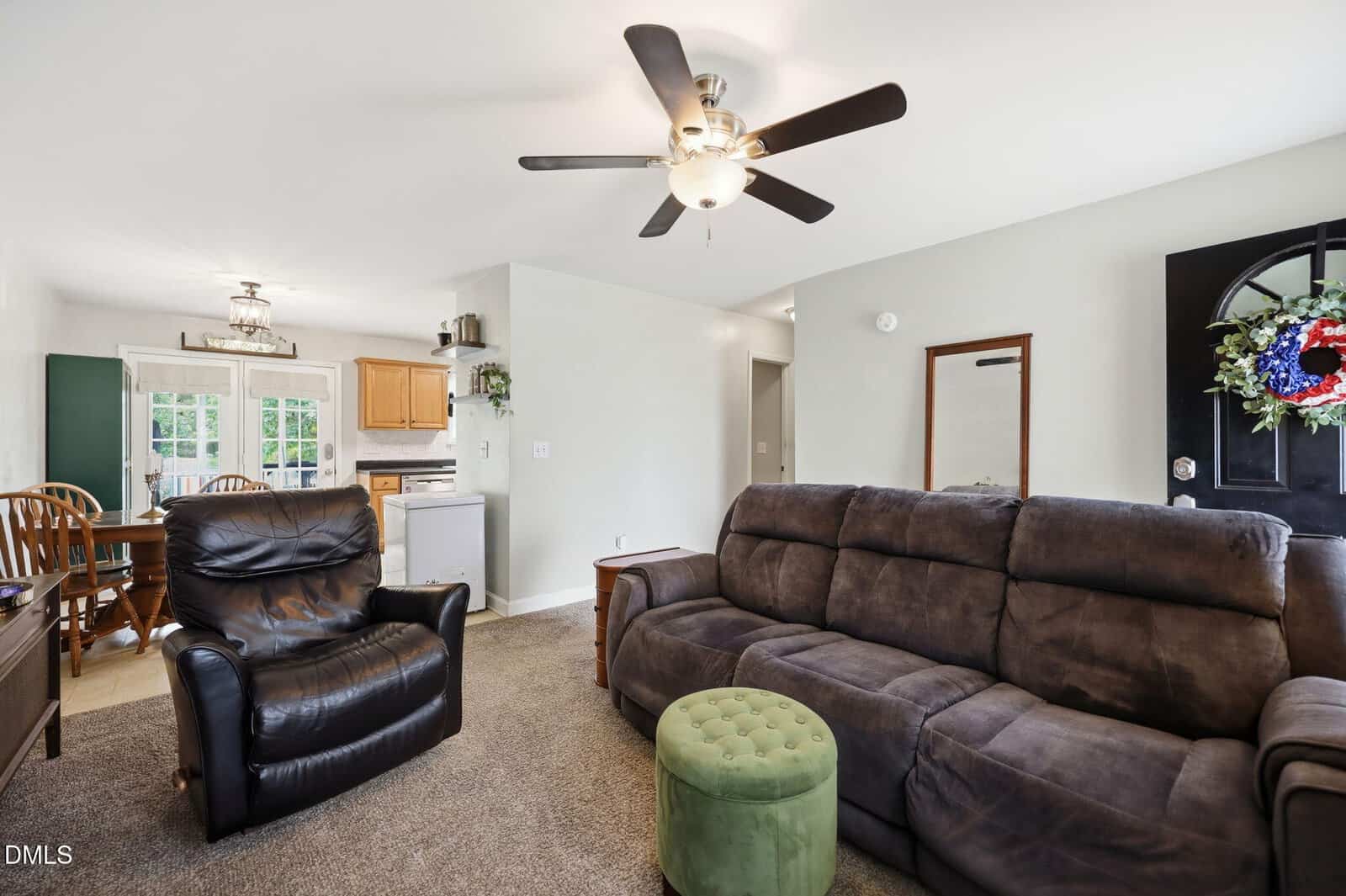 A cozy living room with a dark sofa, black armchair, green ottoman, and ceiling fan. The space connects to a dining area with a wooden table and chairs, and a kitchen with light wood cabinets. A black door features a wreath.