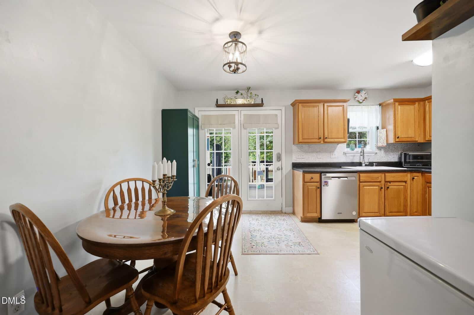 A kitchen with wooden cabinets, stainless steel appliances, a round wooden dining table with four chairs, and double glass doors leading outside. A green cabinet stands against the wall beside the doors.