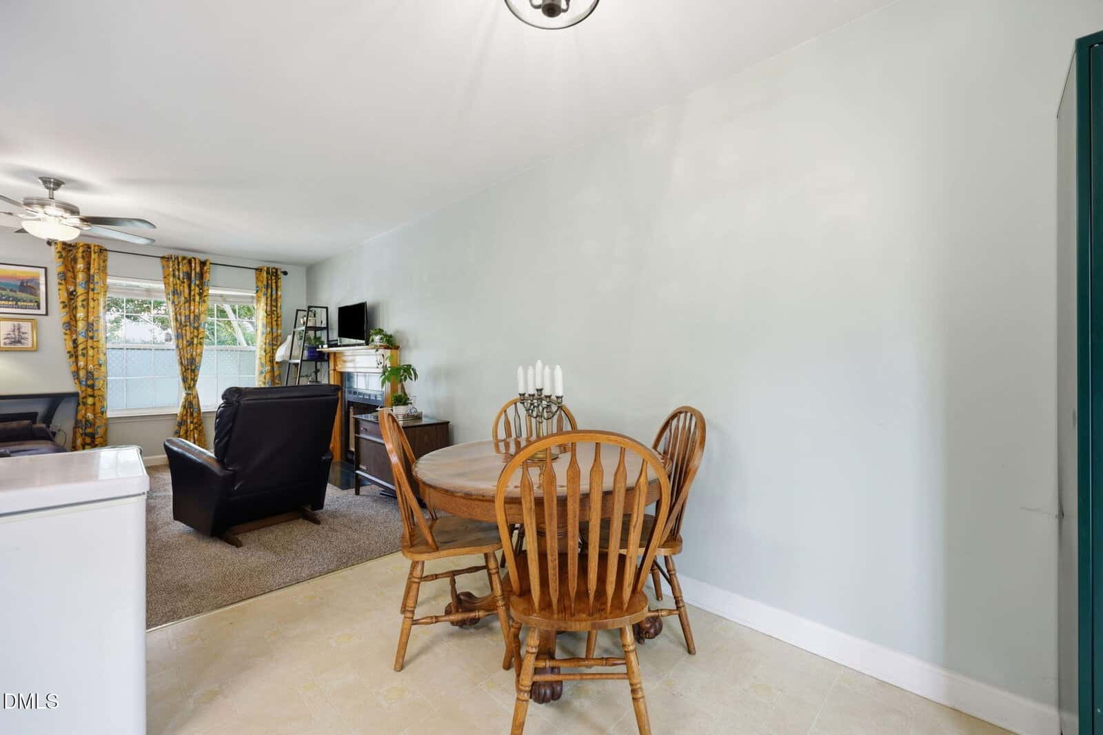 A small dining area with a round wooden table and four chairs is next to a living room with armchairs and yellow curtains. Light green walls and a window let in natural light.
