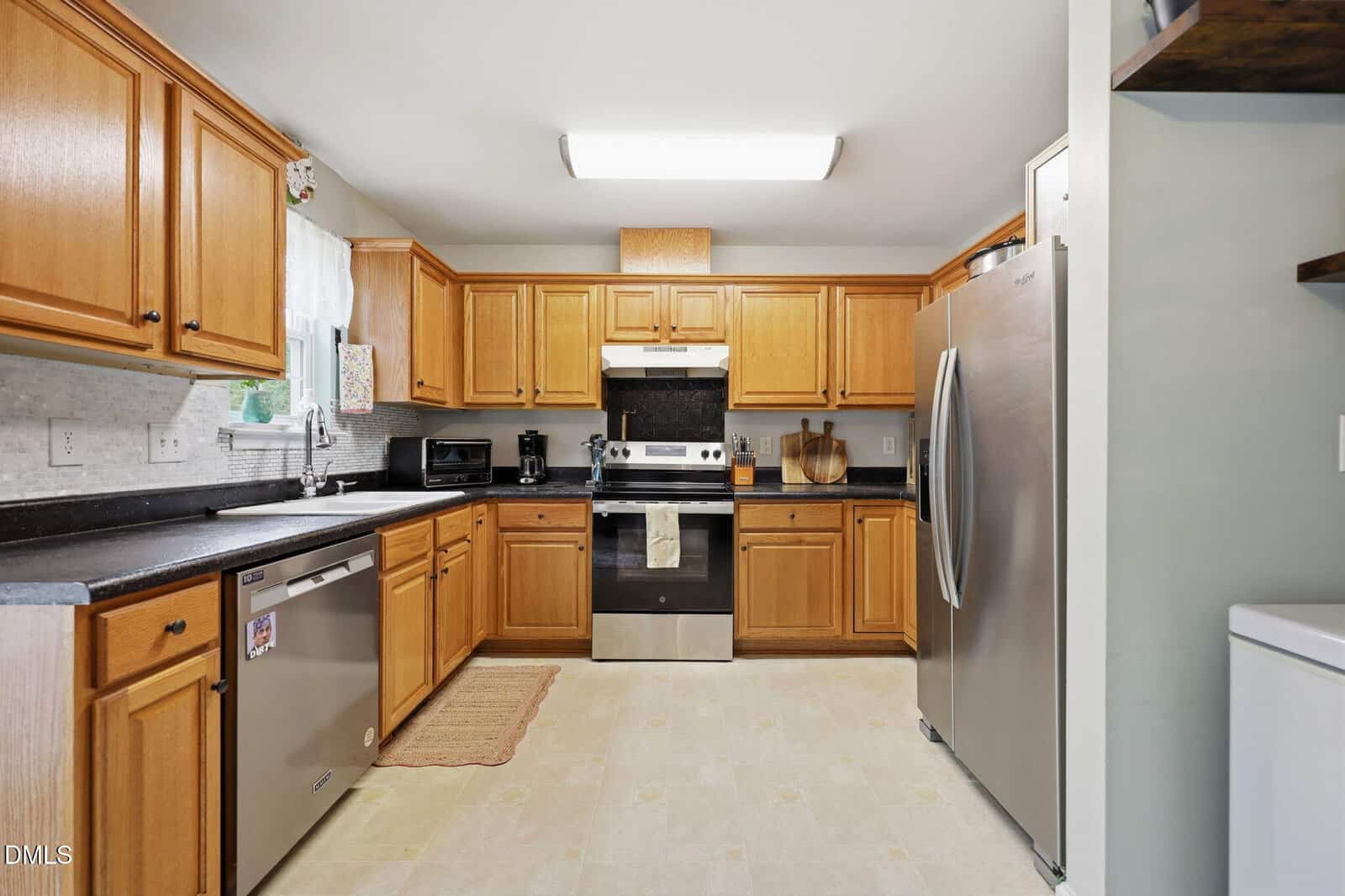 Modern kitchen with light wood cabinets, stainless steel appliances, a dishwasher, oven, refrigerator, black countertops, window over the sink, and beige tiled floor. There are cutting boards and a microwave on the counters.