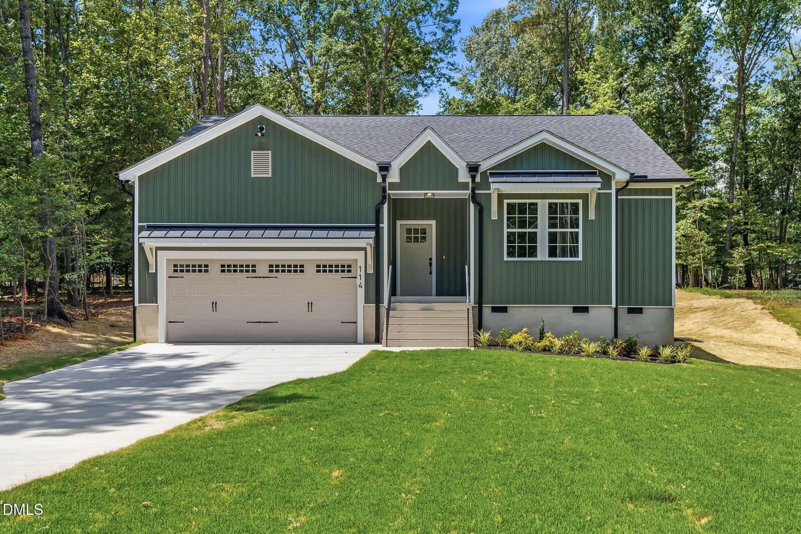 A green single-story house with white trim at 114 Comanche Drive, Louisburg, features a two-car garage, small front porch, and sits on a well-manicured lawn surrounded by tall trees and a concrete driveway.
