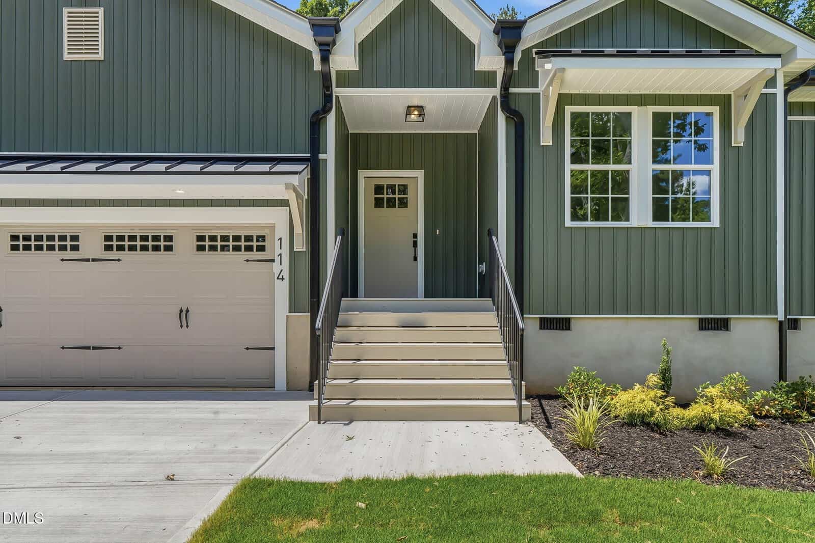 A modern house with green vertical siding, white trim, a white garage door, and stairs with black railings leading to the front door. There are shrubs and mulch along the foundation and a green lawn in front.