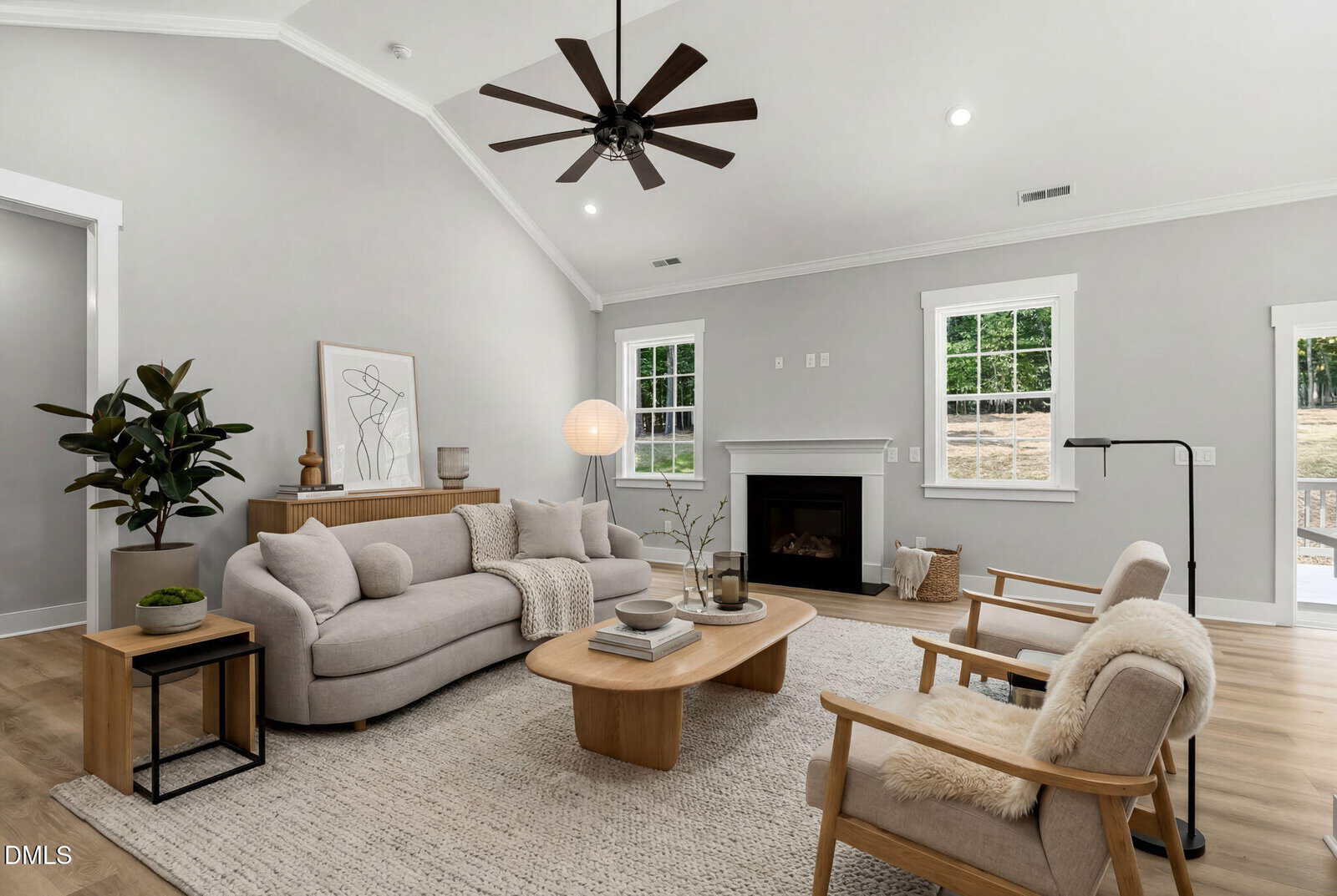 A modern living room with light gray walls, a beige sofa, two armchairs with fur throws, a wooden coffee table, a fireplace, and large windows letting in natural light. Decorative plants and artwork accent the space.