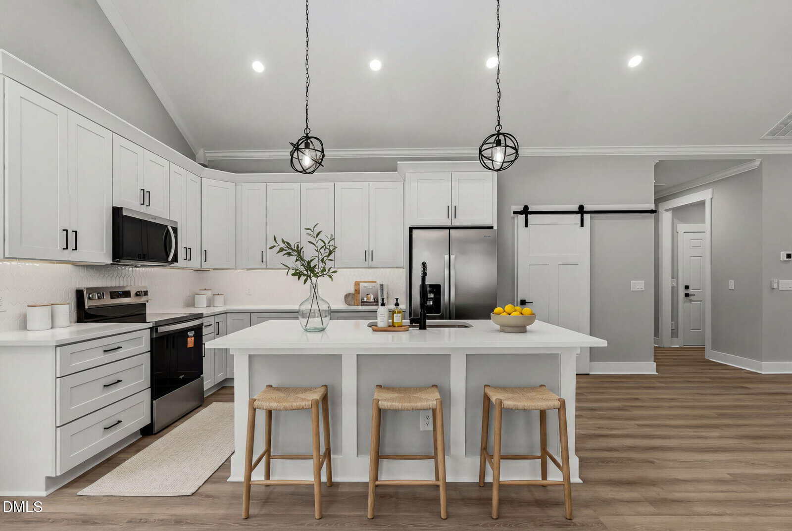 Modern kitchen with white cabinets, stainless steel appliances, a large island with three wooden stools, pendant lights, and light wood flooring. A vase with greenery and a bowl of lemons decorate the island.