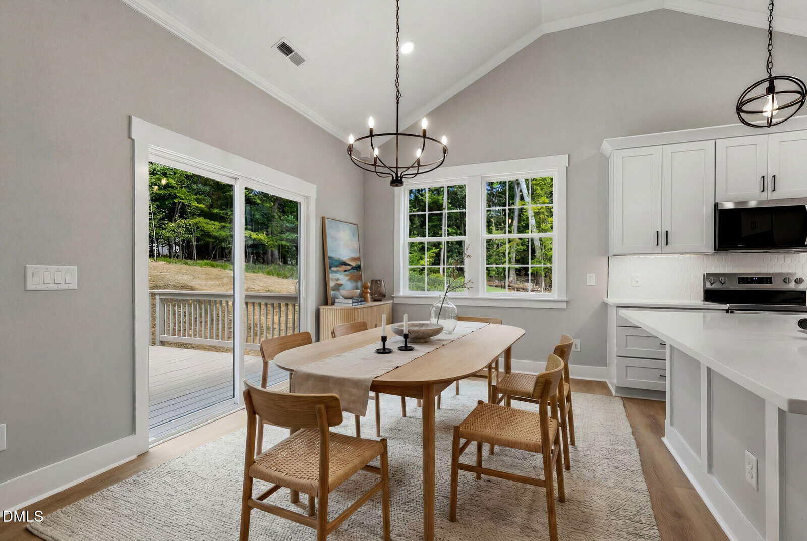A modern dining area with a wooden table, six woven chairs, and two black chandeliers. Large windows and a sliding glass door let in natural light, overlooking a patio and wooded yard. The space has light walls and white cabinets.
