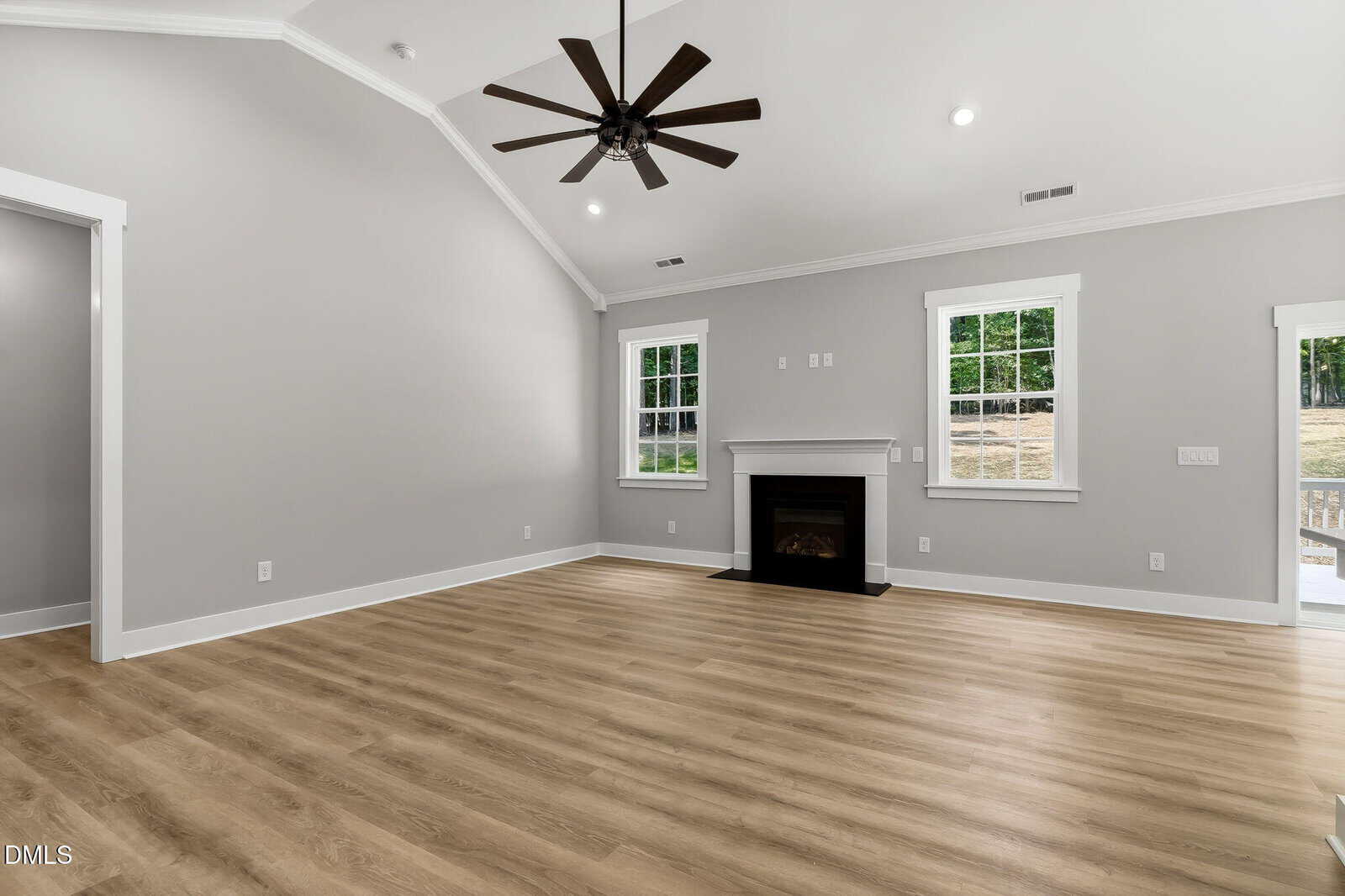 Bright, empty living room with light wood floors, gray walls, white trim, a black fireplace, two windows, and a ceiling fan. Doorway on the right leads outside, and a doorway on the left leads to another room.