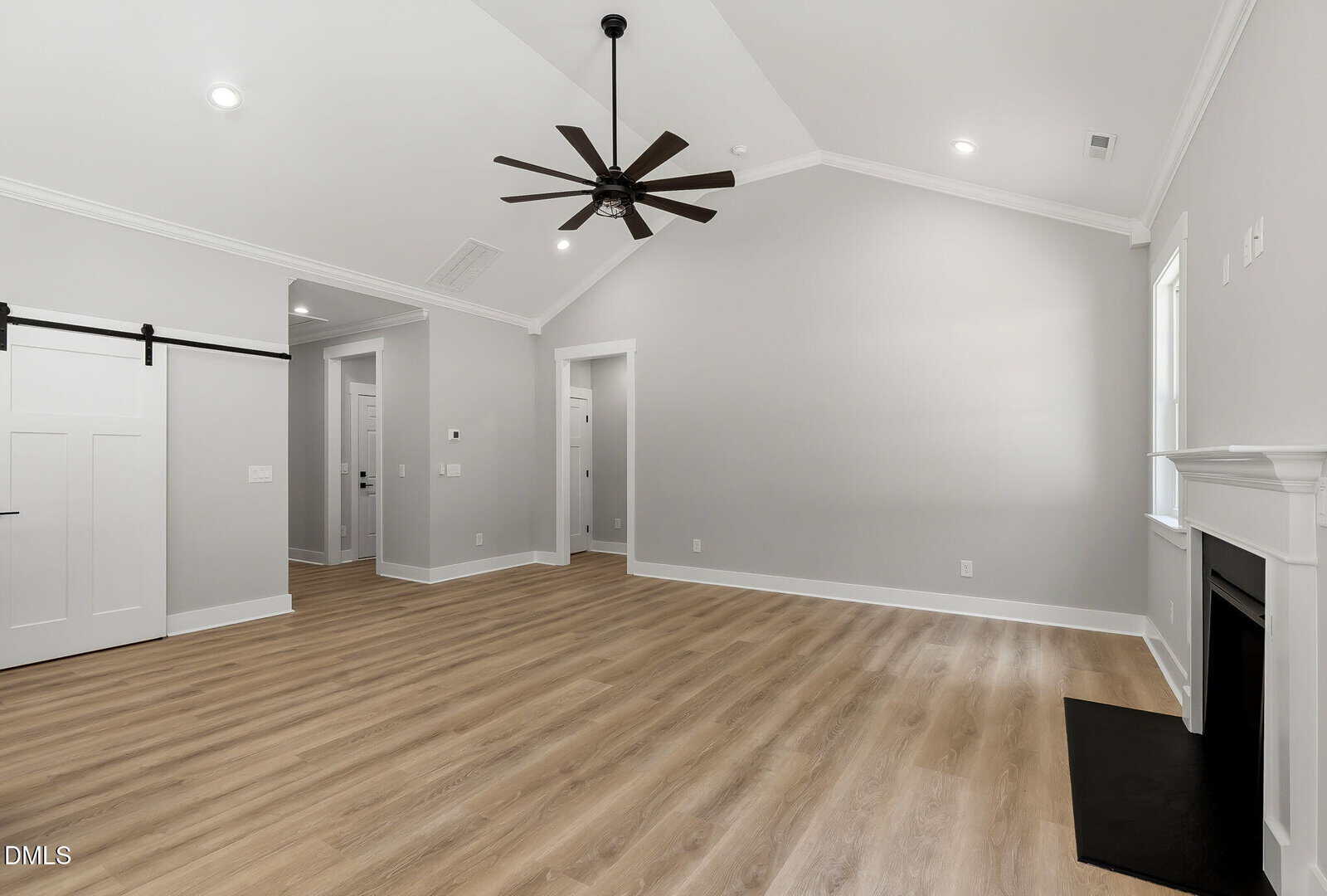 Spacious empty room with light wood floors, gray walls, a large black ceiling fan, white trim, a sliding barn door, and a fireplace on the right side.