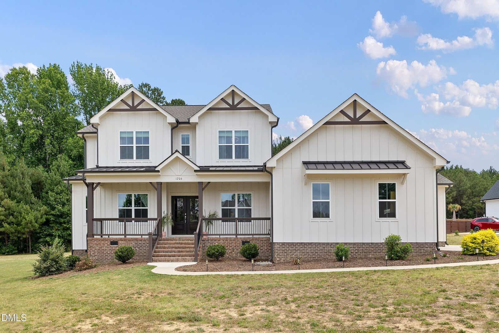 Two-story modern farmhouse at 1206 Pond Side Drive in Creedmoor features white siding, brown trim, a covered front porch, brick foundation, and manicured landscaping on a grassy lawn with trees and blue sky in the background.