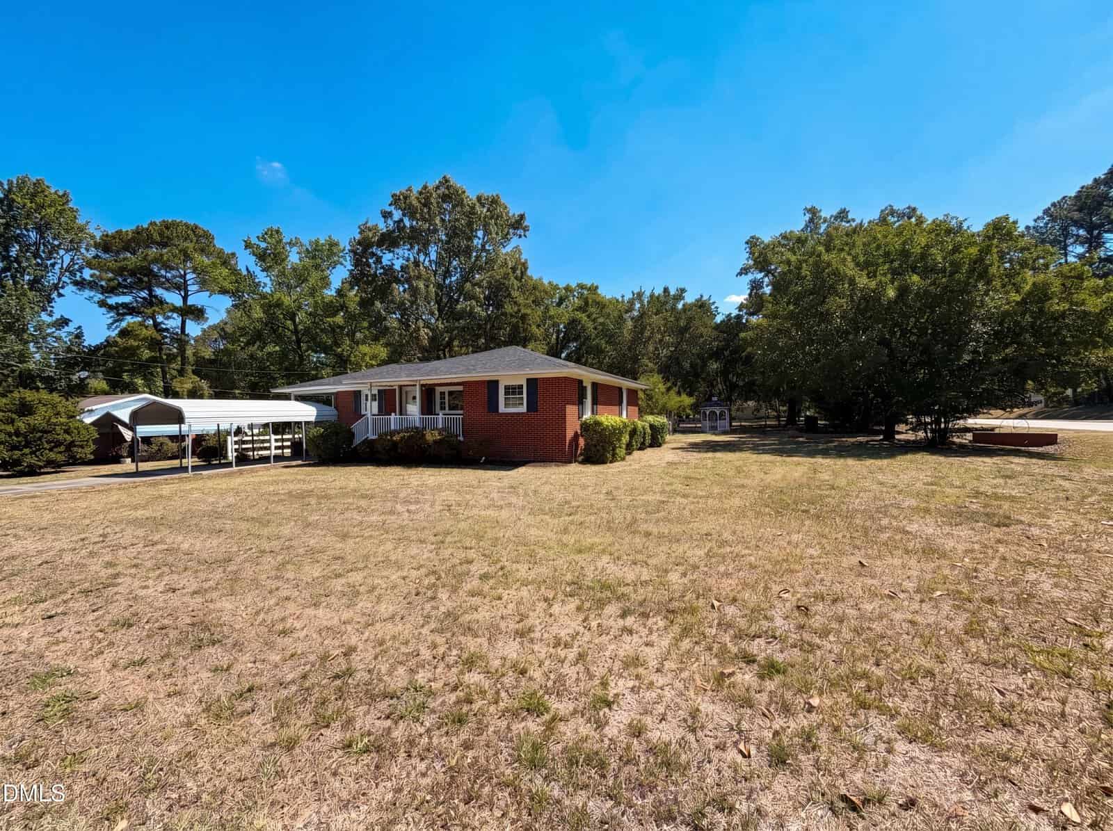 A single-story red brick house with a gray roof sits on a large, dry grassy yard. There is a white carport to the left and several tall trees surrounding the property under a clear blue sky.