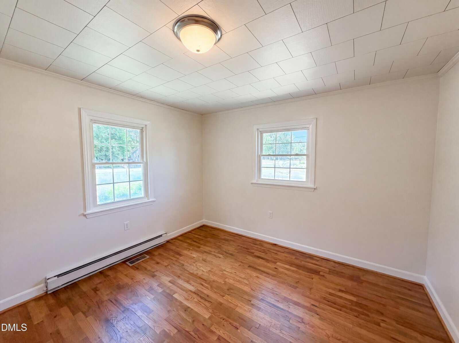 A bright, empty room with hardwood floors, white walls, two windows with views of greenery, a ceiling light fixture, and a white-patterned ceiling.