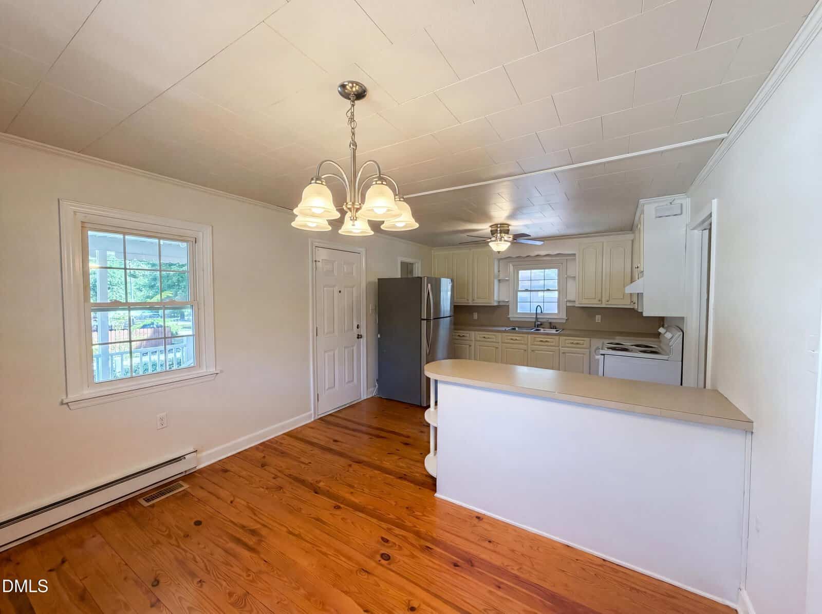 A bright kitchen with wood floors, white cabinets, beige countertops, stainless steel refrigerator, white stove, and a central light fixture. A window and door are on the left, letting in natural light.