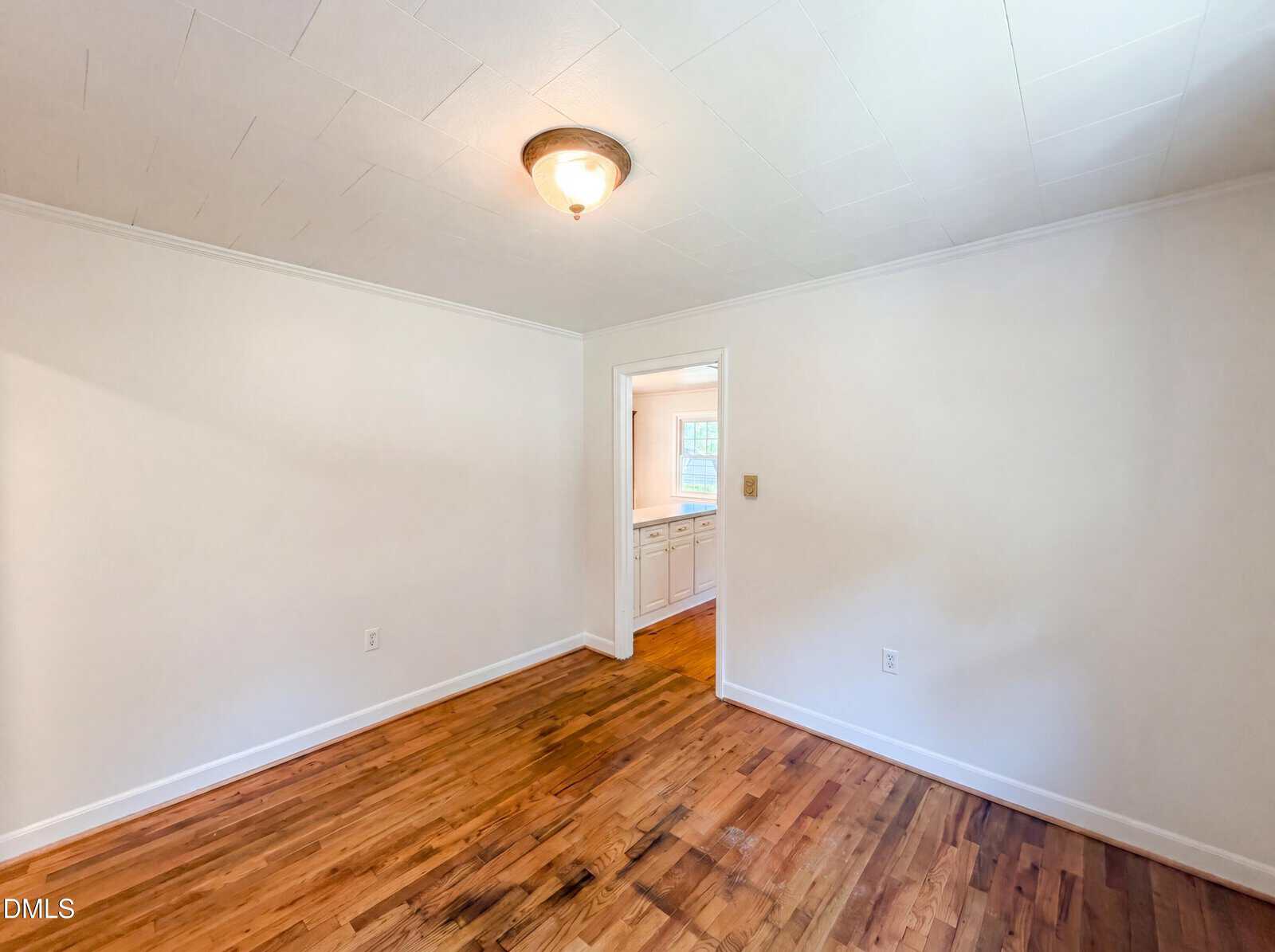 Empty room with white walls, wood flooring, a ceiling light, and an open doorway leading to a kitchen with white cabinets and a window.
