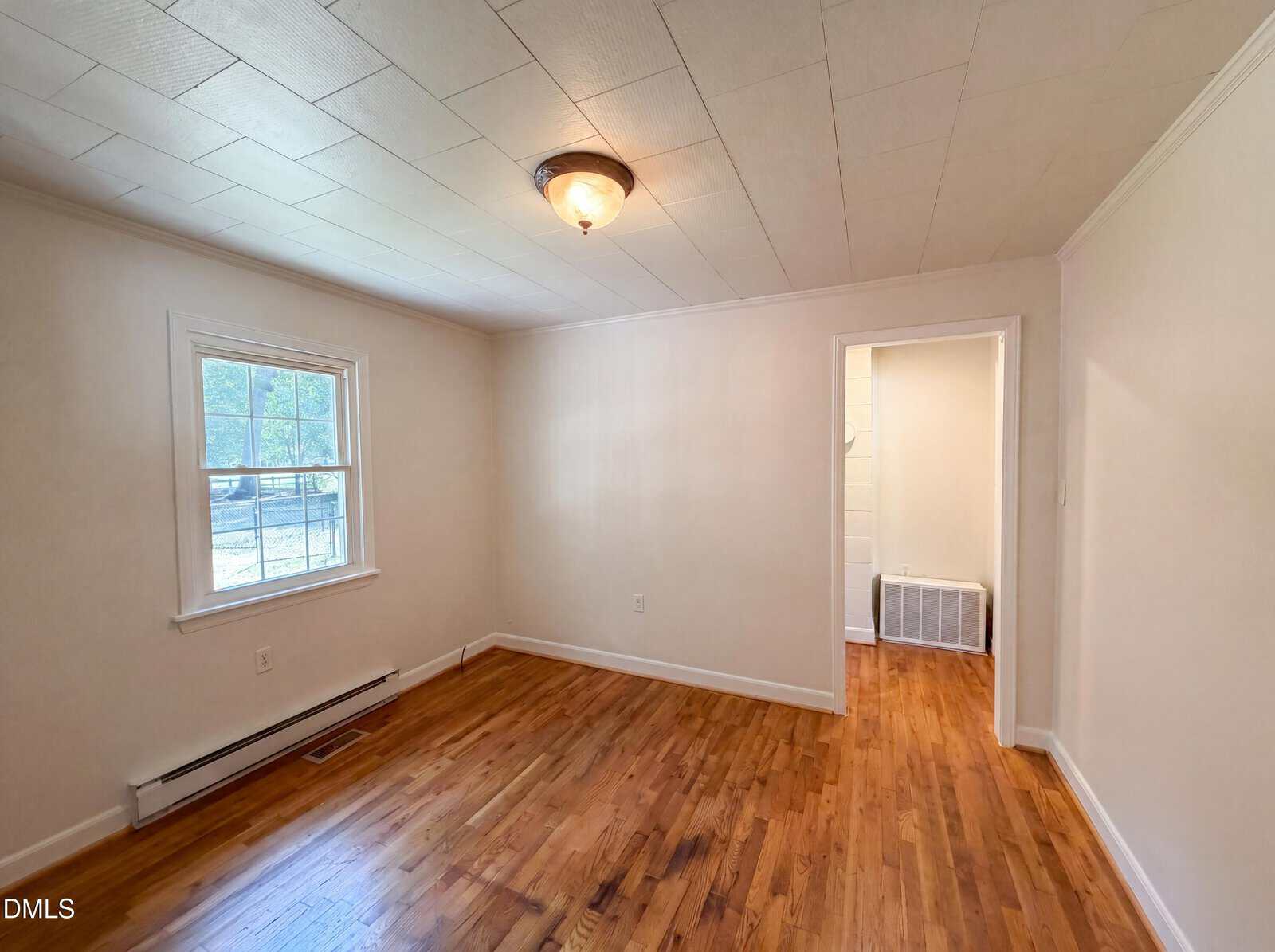 Empty room with hardwood flooring, white walls and ceiling, a single window on the left, ceiling light fixture, and an open doorway leading to another area with more hardwood floors.