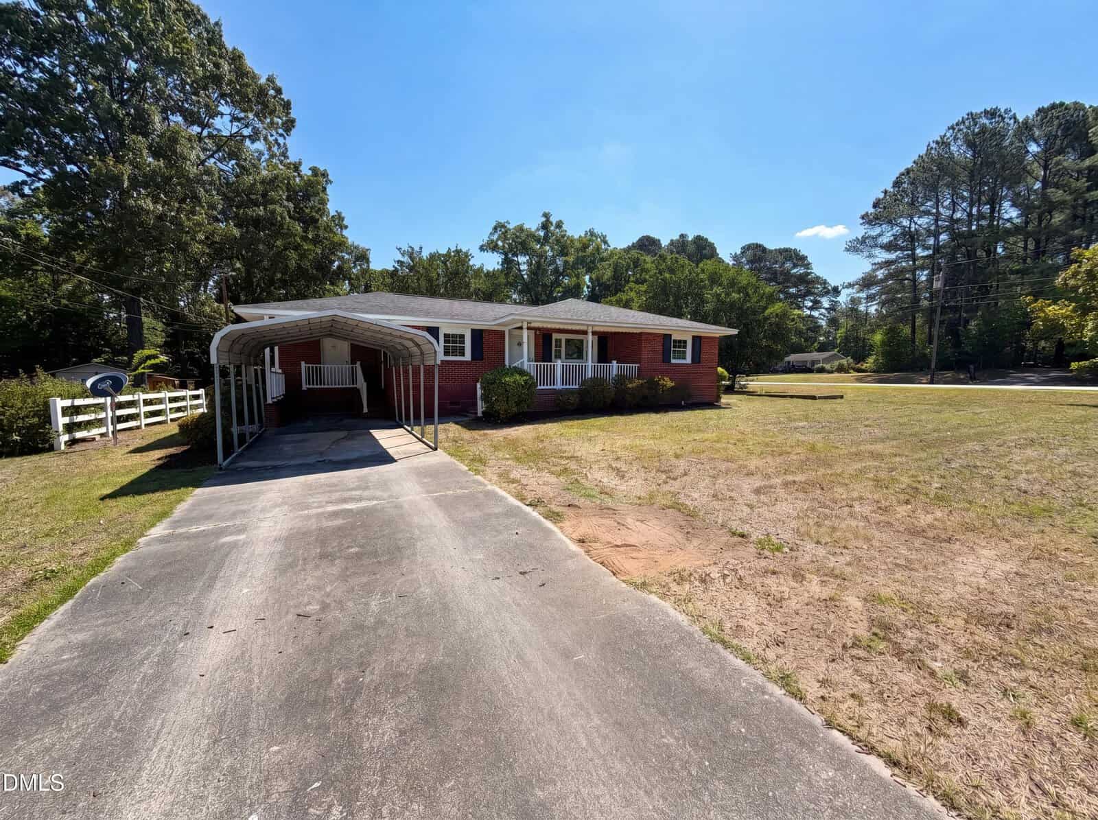 Charming single-story red brick house at 300 7th Street, Butner, with a covered carport, welcoming front porch, large grassy yard, and paved driveway. Mature trees surround the property on a sunny day.