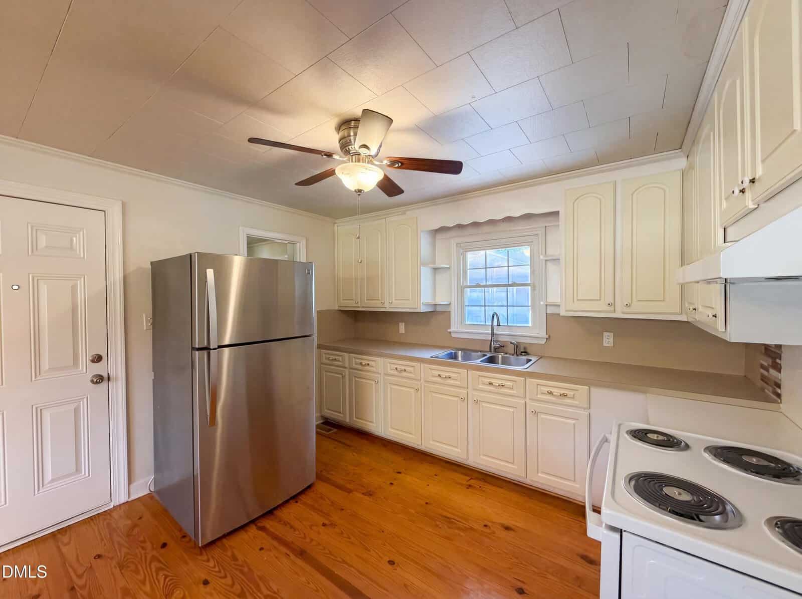 A bright kitchen with white cabinets, stainless steel refrigerator, electric stove, ceiling fan with light, wood floor, and a window above the sink letting in natural light.