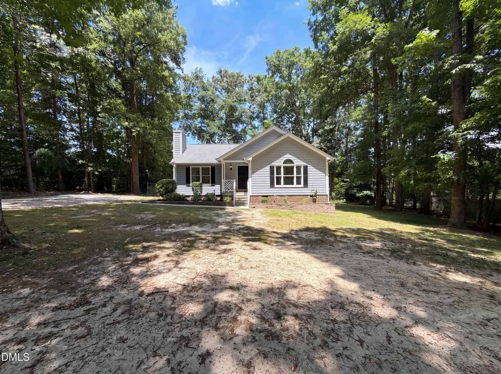 A small, single-story gray house with white trim sits among tall trees at 328 Thornwood Lane in Youngsville. A large dirt and patchy grass front yard stretches out beneath a blue sky, with tree shadows covering parts of the yard and driveway.