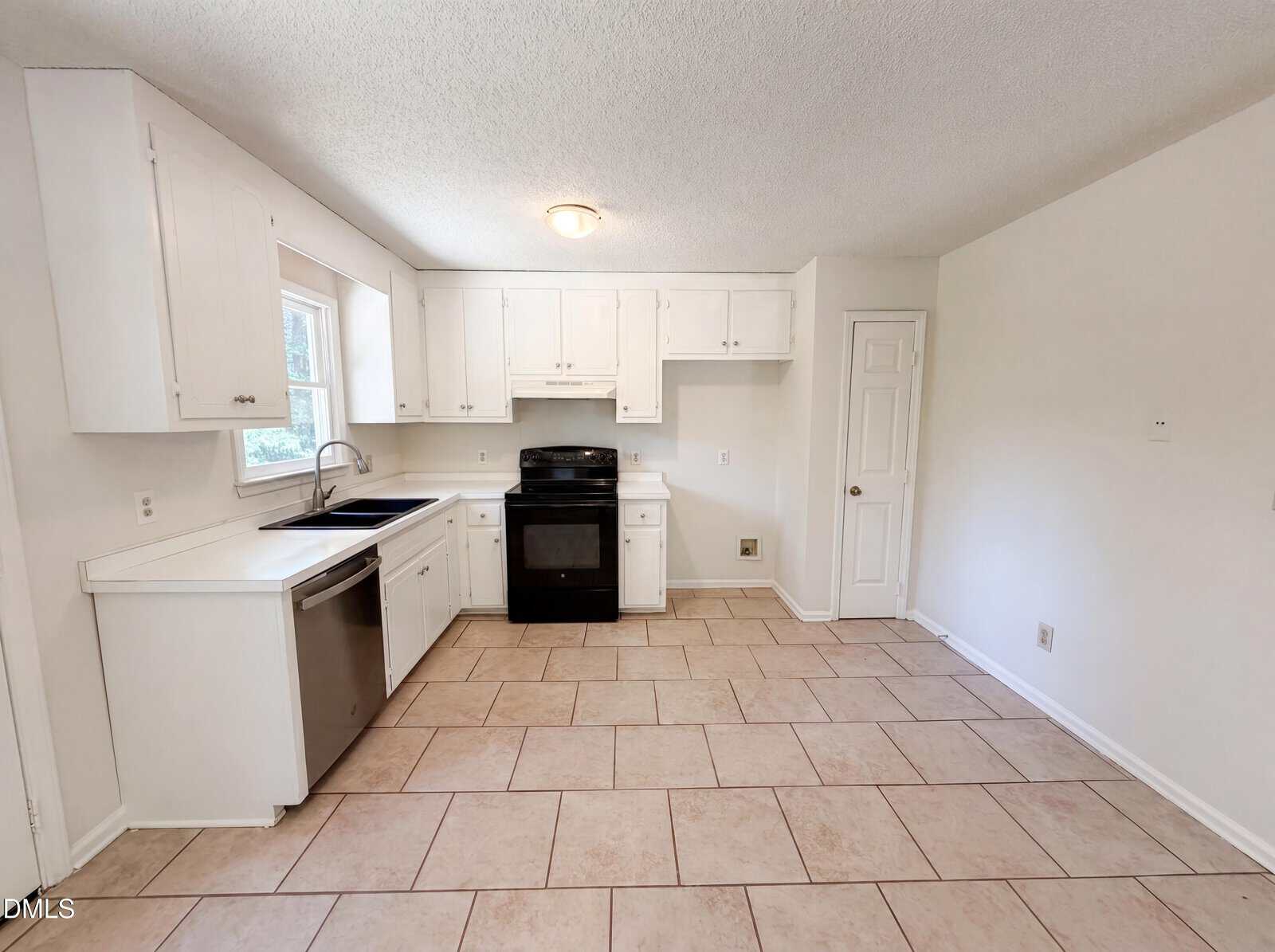 Bright kitchen with white cabinets, black stove and oven, stainless steel dishwasher, single sink under a window, tiled floor, and a white door on the right wall. Walls are painted white and the space is empty.